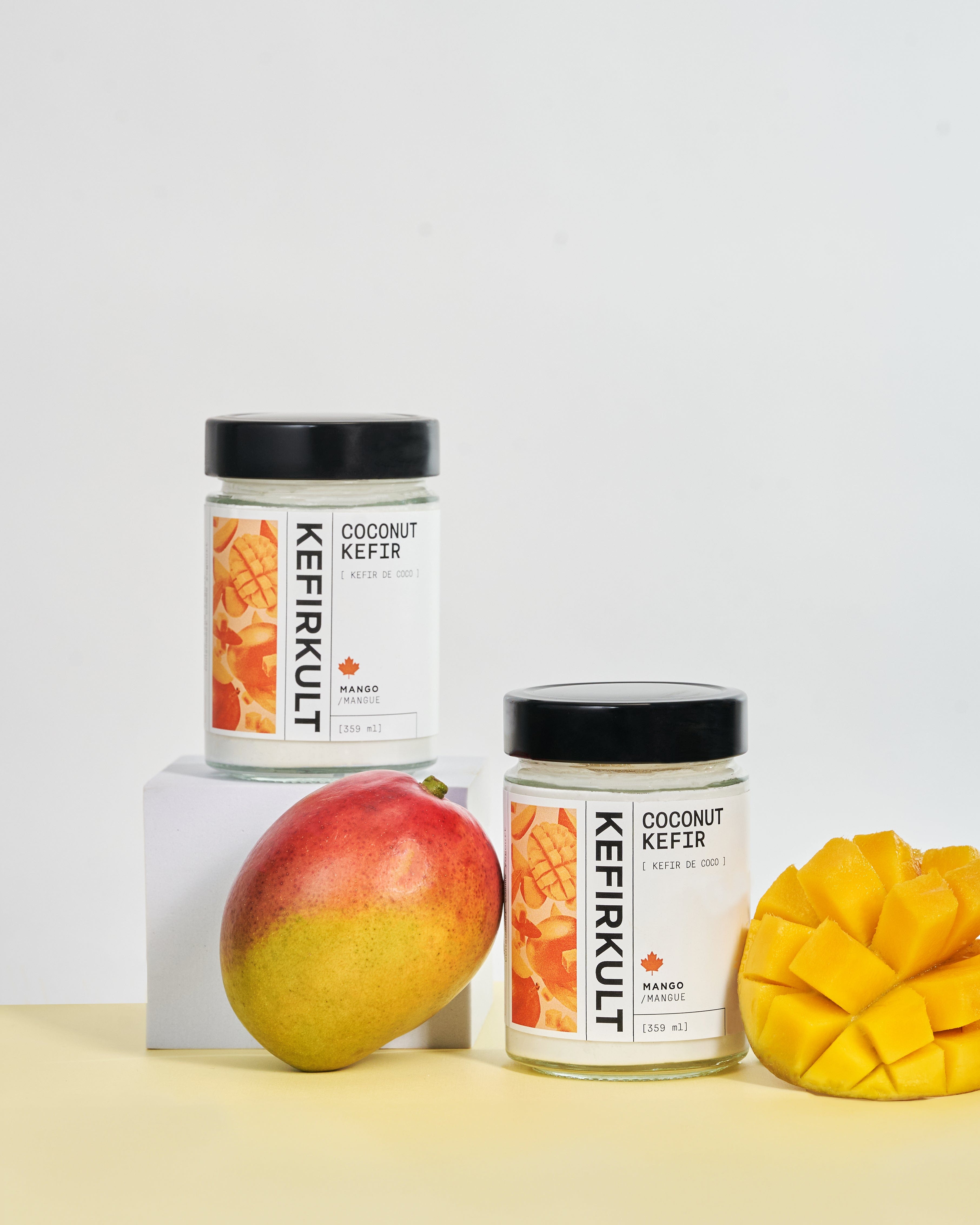 Two jars of KefirKult coconut kefir, label text: KEFIRKULT COCONUT KEFIR KEFIR DE COCO MANGO MANGUE 359 ml; whole and sliced mango.