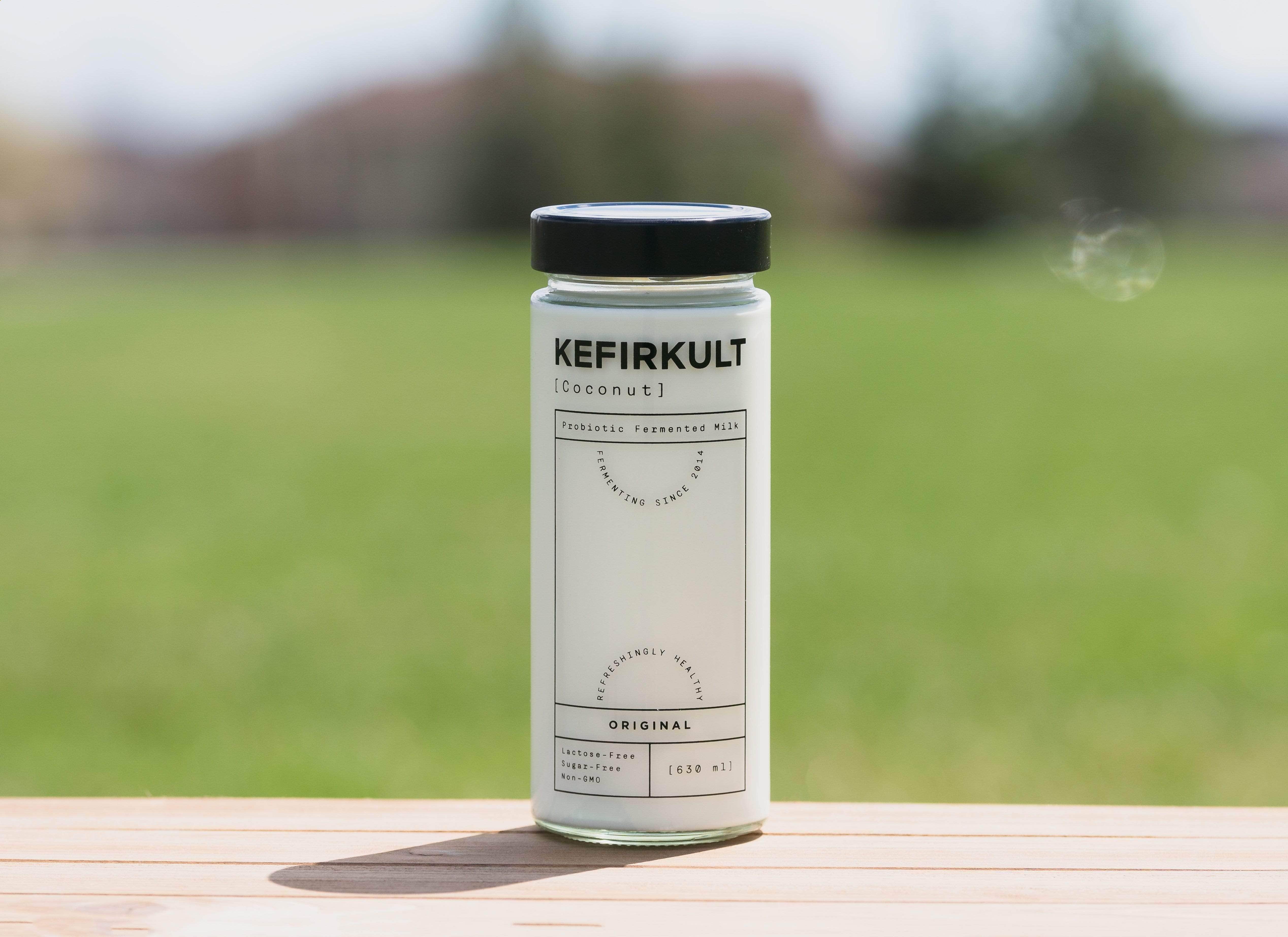 Bottle of Kefirkult coconut probiotic fermented milk with a black lid sits on a wooden surface outdoors, with a blurred green field in the background.